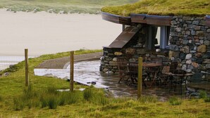 A stone building with a grassy roof overlooking a lake, featuring a patio with chairs and a table.
