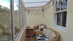 A cozy room with a wicker sofa, a small table, and a lamp. There are bunting decorations on the wall and a window overlooking a stone wall. - 2 bedroom accommodation in Alnwick (Alnwick)
