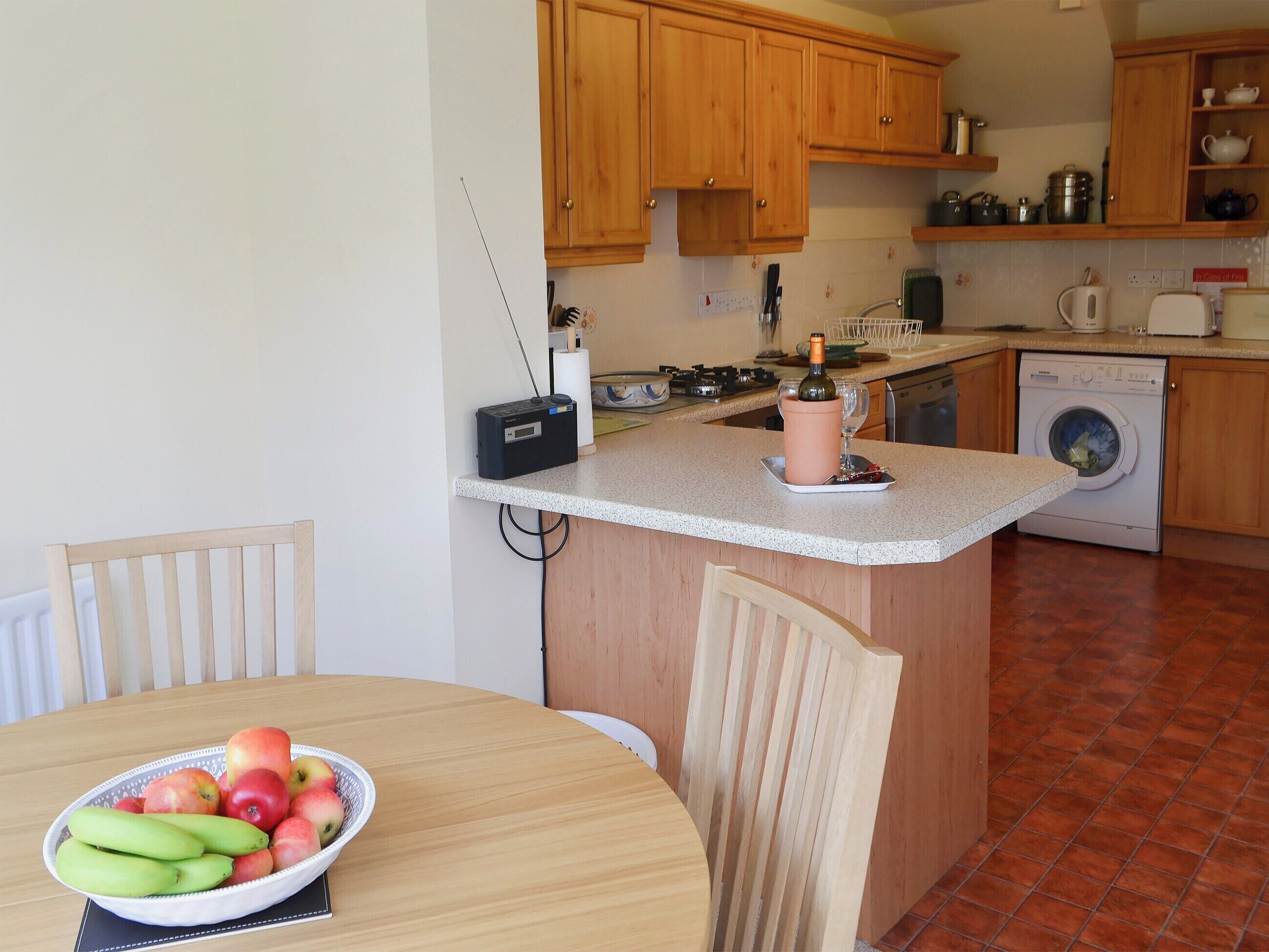 A kitchen with wooden cabinets, a washing machine, a radio, and a table with a bowl of fruit.