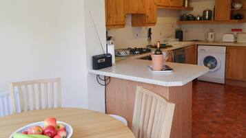 A kitchen with wooden cabinets, a washing machine, a radio, and a table with a bowl of fruit.