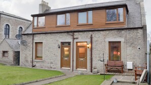 A two-story stone building with numbered doors, a wooden upper floor, and a small front yard with a bench.