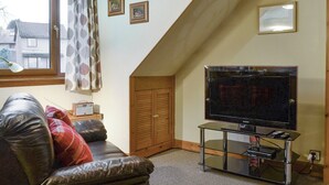 A living room with a leather sofa, a flat-screen TV on a glass stand, a window with curtains, and a wooden door.