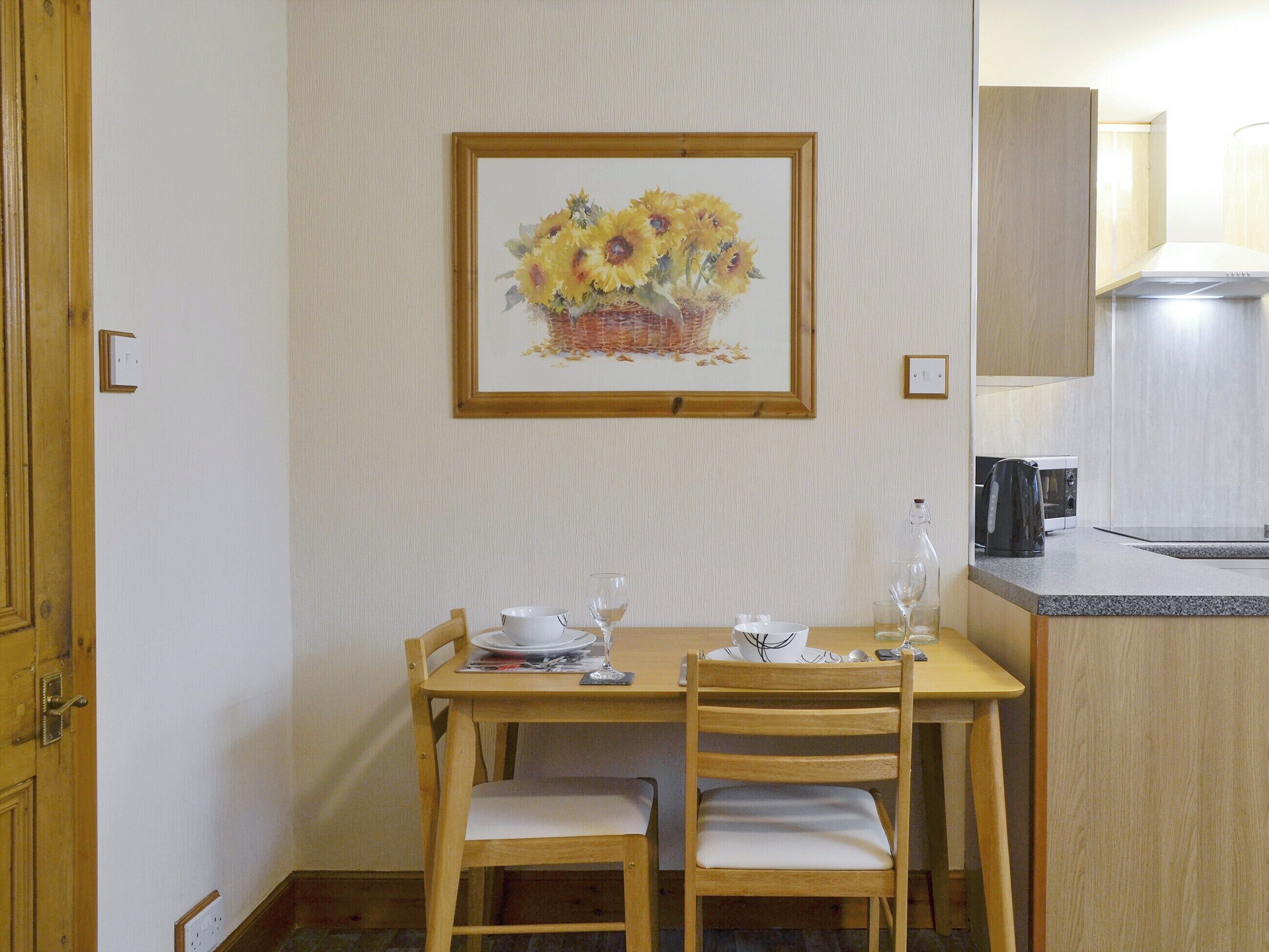 Uma área de refeições com uma mesa de madeira, duas cadeiras, um quadro emoldurado com girassóis e um balcão de cozinha com uma chaleira e um micro-ondas.