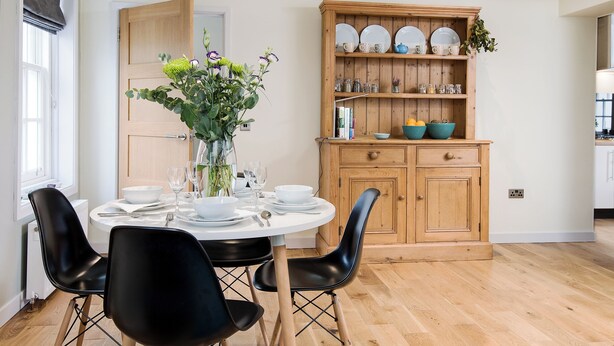 Zona pranzo con tavolo rotondo, sedie nere, una credenza in legno con stoviglie e un vaso con fiori.