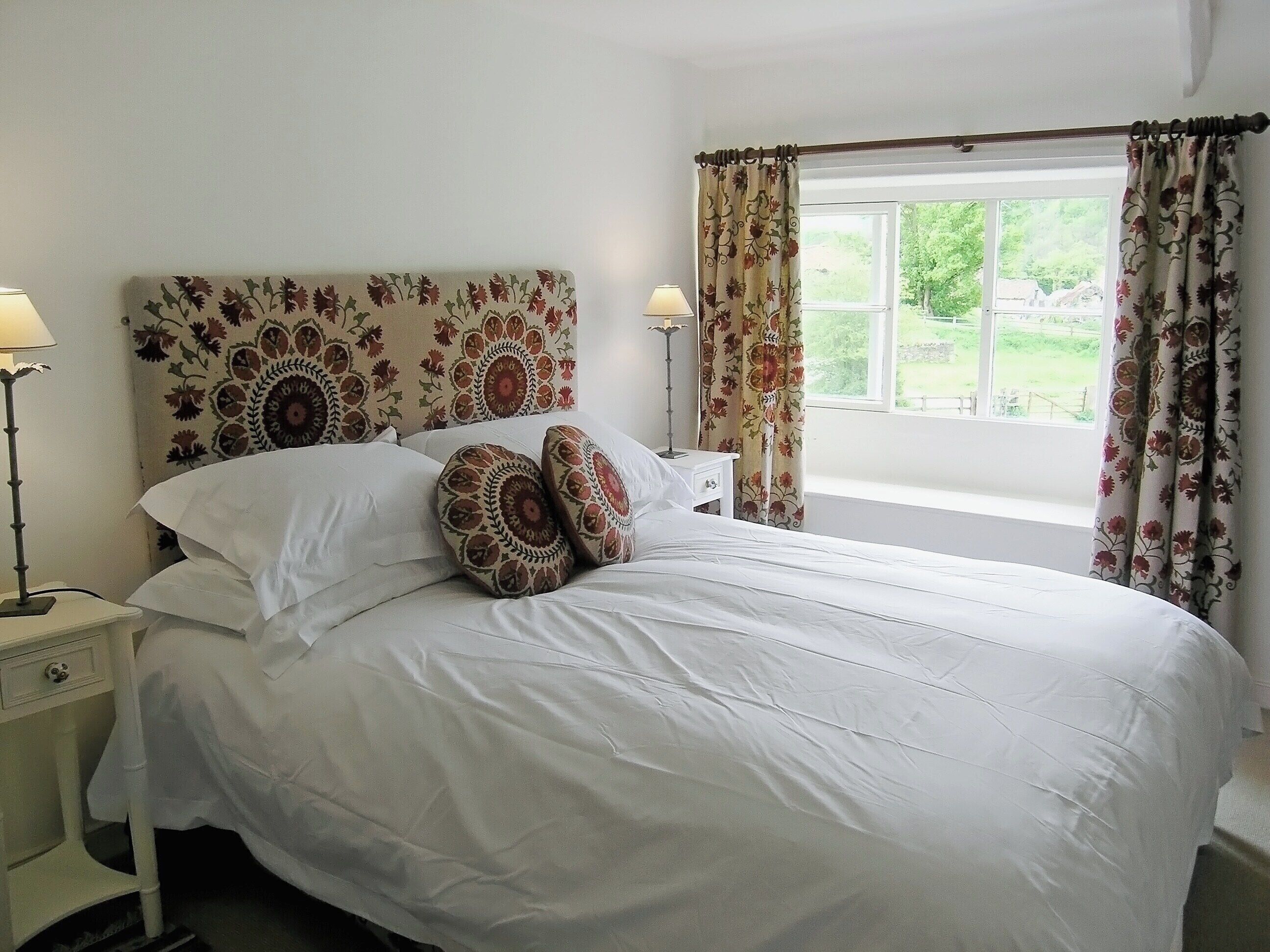 A neatly made bed with white linens and patterned pillows, a bedside table with a lamp, and a window with floral curtains.