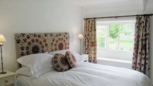 Een netjes opgemaakt bed met witte lakens en gestreepte kussens, een nachtkastje met een lamp, en een raam met bloemen gordijnen.