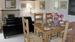 Een eethoek met een houten tafel gedekt voor vier personen, omringd door houten stoelen met gevlochten zitplaatsen. Een zwarte leren bank staat links, en er is een open haard zichtbaar op de achtergrond.