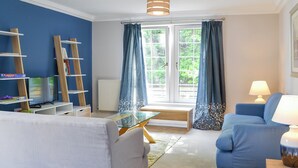 A living room with a blue sofa, a glass coffee table, and a TV. There are bookshelves and a ladder. The room has a window with curtains and a lamp. - 3 bedroom accommodation in Edinburgh (Edinburgh)