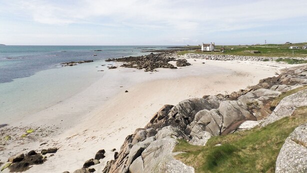 A coastal landscape with a sandy beach, rocky outcrops, and a white building in the distance.