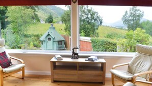 Una stanza con un'ampia finestra vista su un paesaggio verde, un tavolo di legno con libri e una figurina, e due sedie.