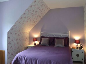 Een slaapkamer met een schuine plafond, een bed met een paarse sprei, twee nachtkastjes met lampen, en bloemenbehang.