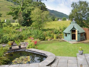A garden with a pond, a small wooden structure, and a grassy area with trees and hills in the background. - 6 bedroom accommodation in Ballachulish, near Fort William (Ballachulish, near Fort William)