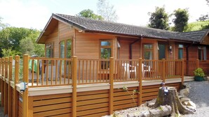 A wooden cabin with a deck, large windows, and a gravel driveway.