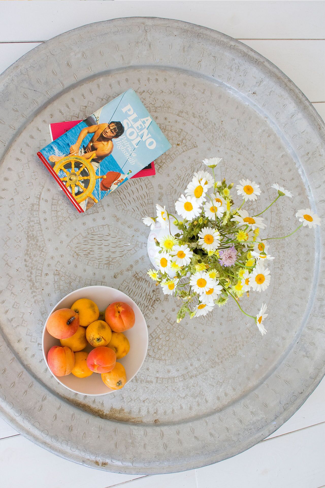 A round ceramic tray with a floral pattern, a bowl of peaches, a bouquet of daisies, and a magazine titled "AENIO PLENO".