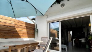 Una terraza de madera con una mesa, sillas y un gran paraguas, además de un ventanal corredero que da a un espacio interior.
