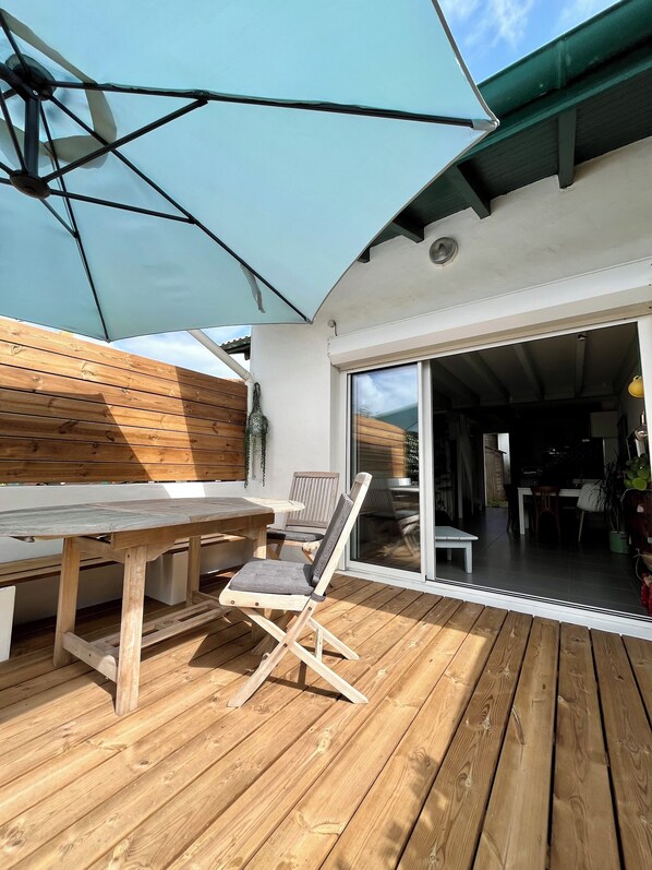 A wooden deck with a table and chairs, a large umbrella, and a sliding glass door leading to an interior space.