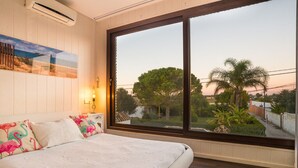 Une chambre avec une grande fenêtre, un lit agrémenté d’oreillers à motifs floraux, un climatiseur fixé au mur et un tableau représentant une scène de plage.