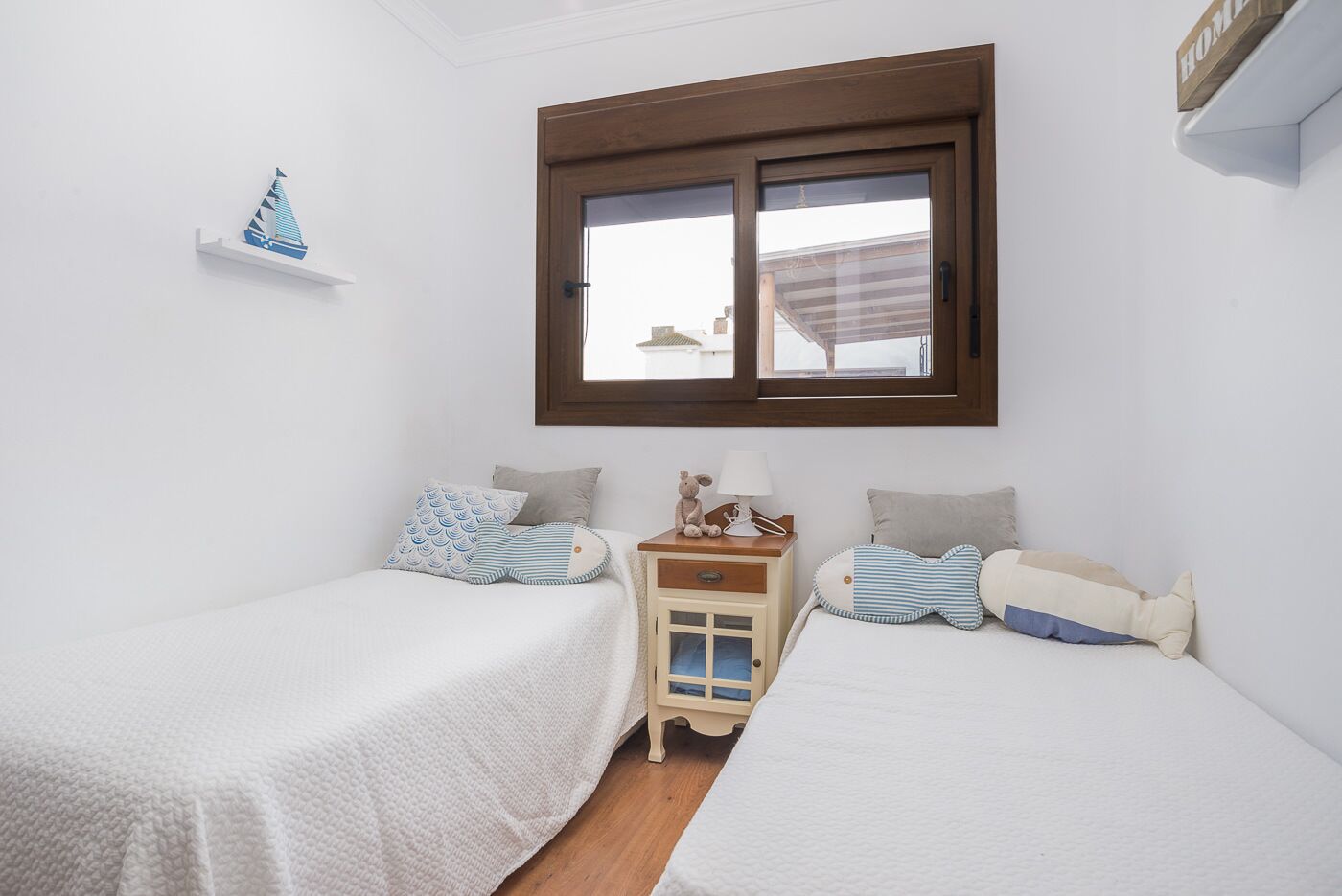 A bedroom with two single beds, a nightstand, a small shelf with a decorative item, and a window with a view of buildings outside.