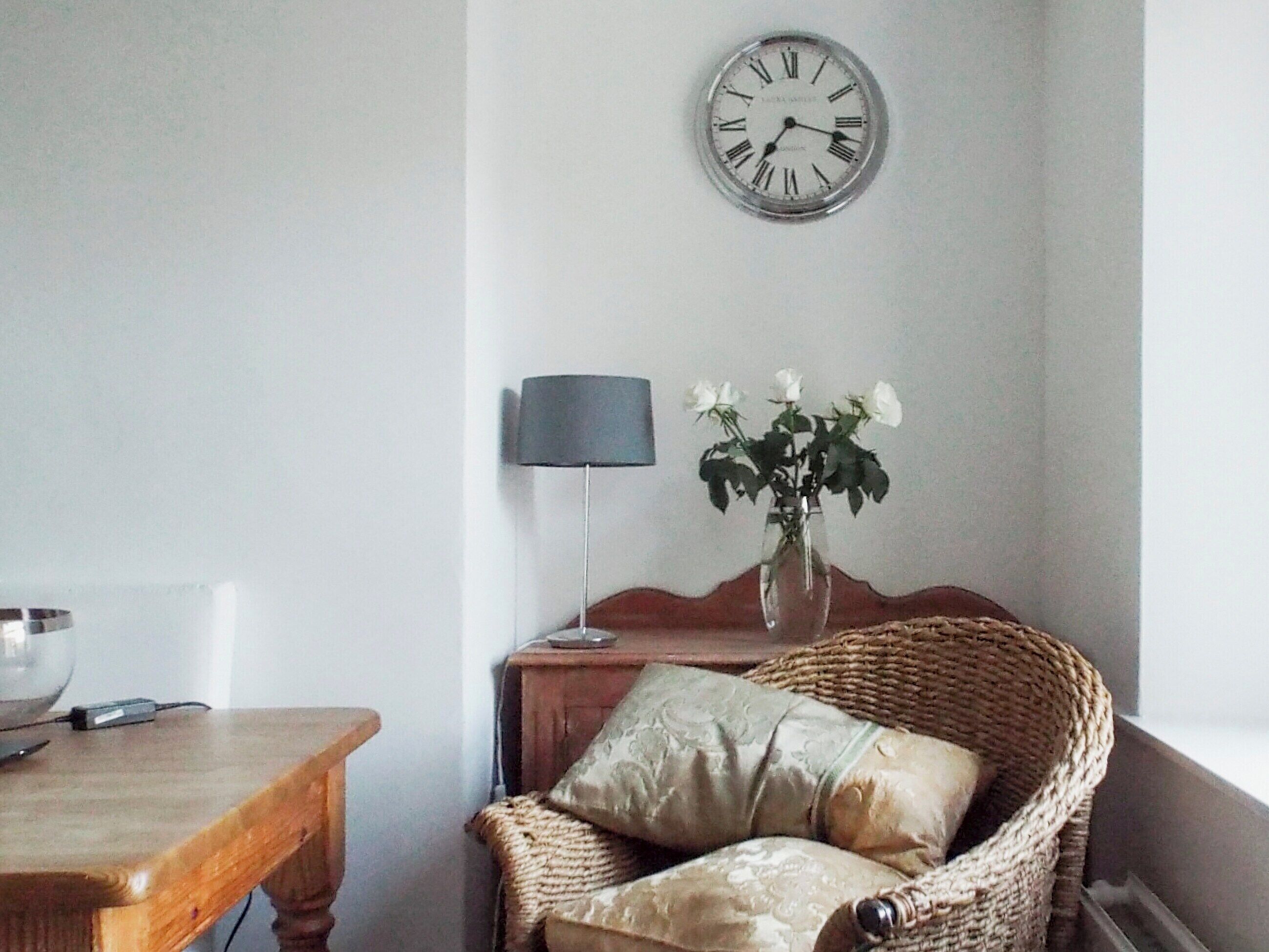 A room with a wooden table, a wicker chair with cushions, a vase with white flowers, a wall clock, and a lamp.