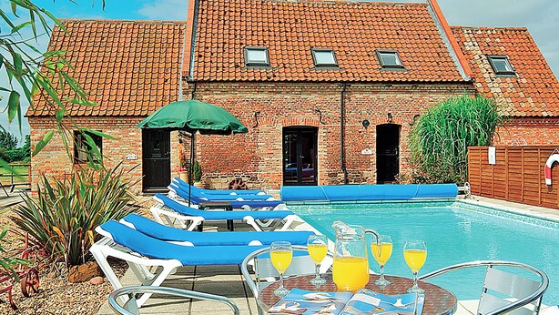 A poolside area with lounge chairs, a table with drinks, and a building with a tiled roof. - Timbers Barn - E3863 (Runcton Holme)