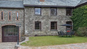 A stone building with a brick chimney, a large arched door, and a small patio with outdoor furniture. - Bridge House - UKC1340 (Heasley Mill, near South Molton)