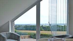 A bedroom with a large window, a bed with towels, a chair, and a view of a field and buildings.