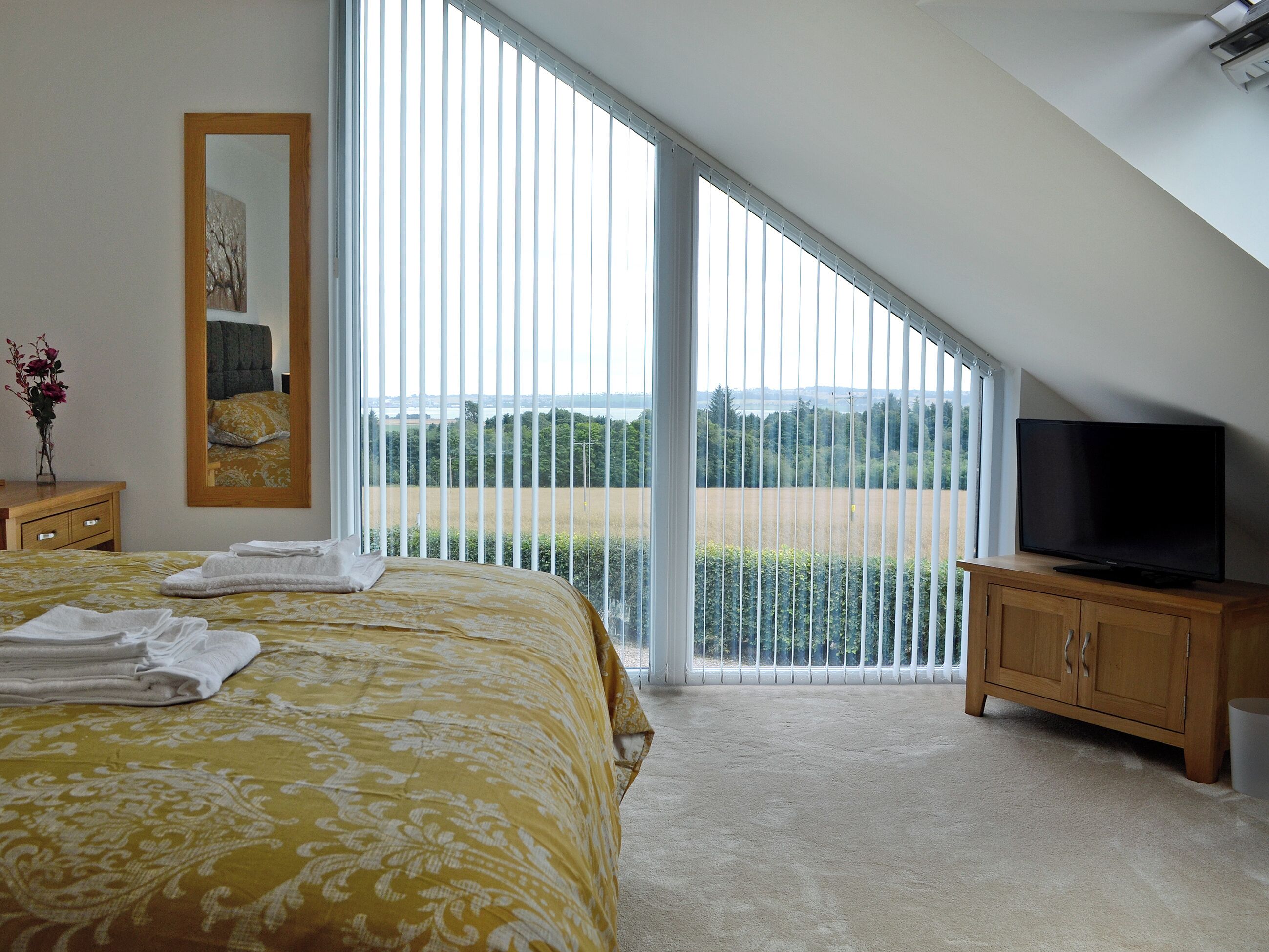 Ein Schlafzimmer mit Bett, Fernseher, Spiegel und Blick auf ein Feld durch große Fenster.