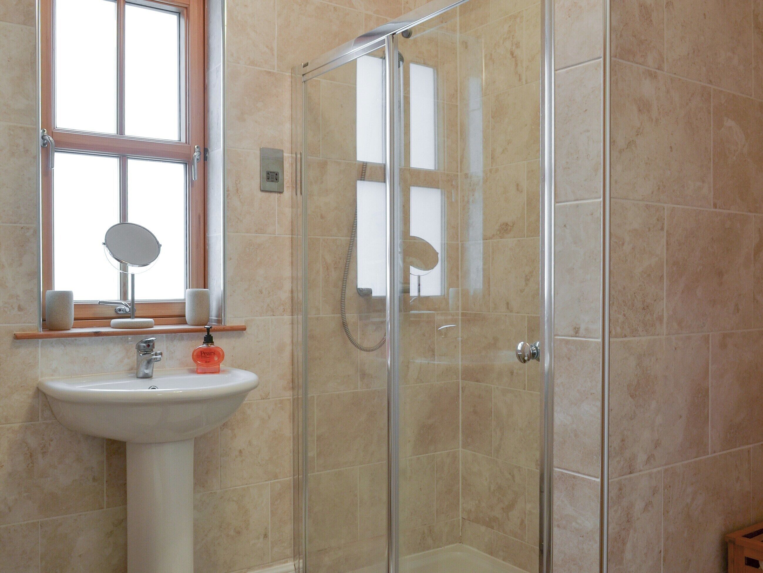 Een badkamer met een douchecabine van glas, een wastafel op een voetstuk en een raam boven de wastafel.