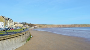 A coastal town with a sandy beach, a promenade lined with parked cars, and buildings along the shore.