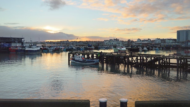 A harbor with boats docked at piers, a city skyline in the background, and a sunset over the water.