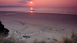 A beach with a sunset, calm sea, and distant mountains. - 2 bedroom accommodation in Godolphin Cross, Helston (Godolphin Cross, Helston)
