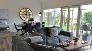 En moderne stue med en krogsofa, et spiseområde med bord og stole, et stort vægur og udsigt til det fri gennem store vinduer.