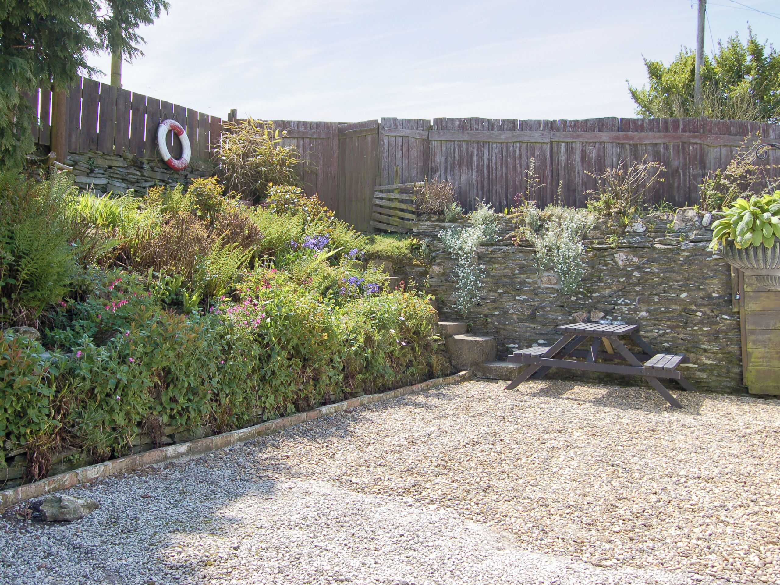 A gravel patio with a wooden table and benches, surrounded by a garden with various plants and a wooden fence.