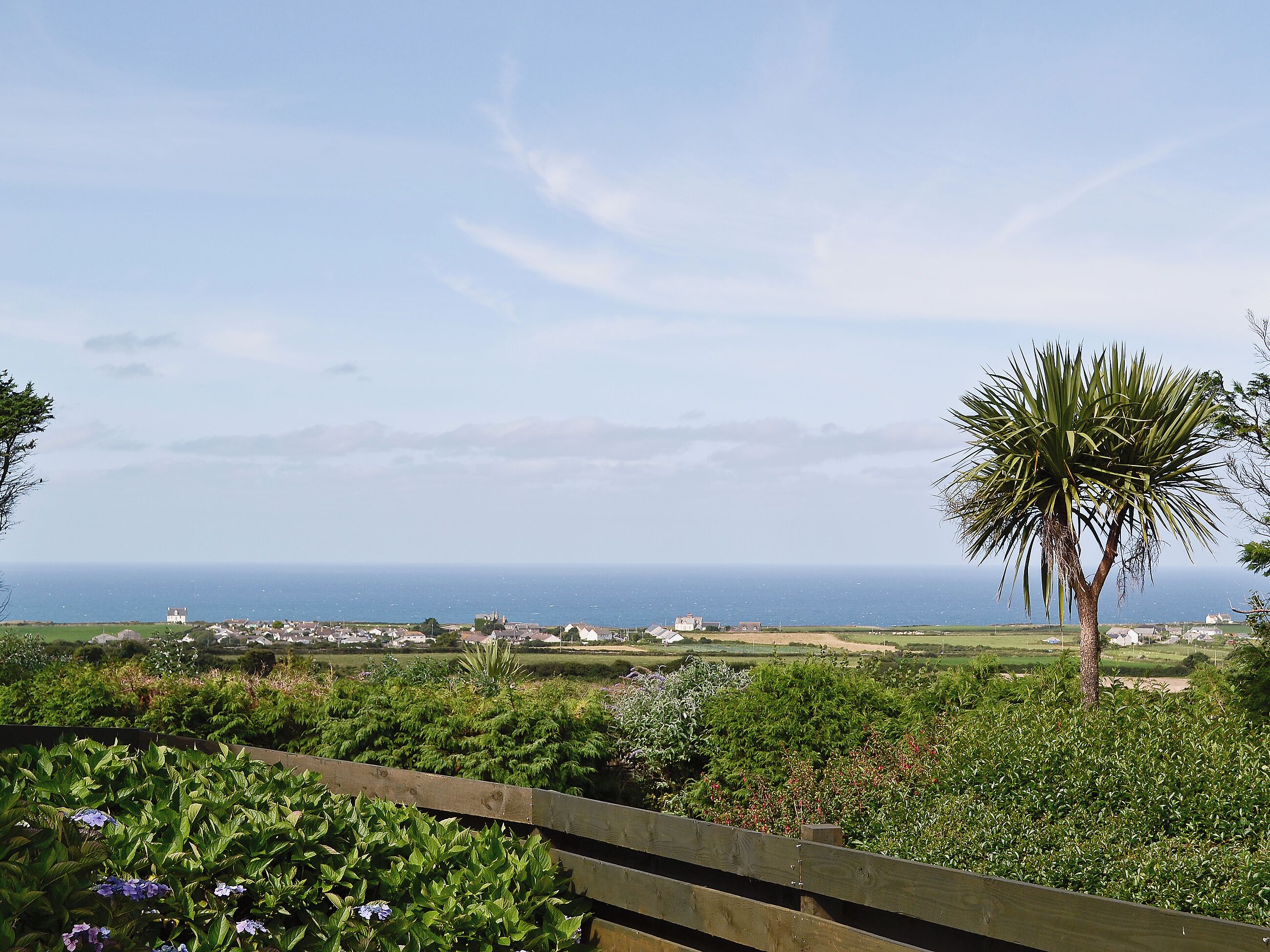 A coastal landscape with a wooden fence, a variety of greenery, and a distant view of the sea and buildings.