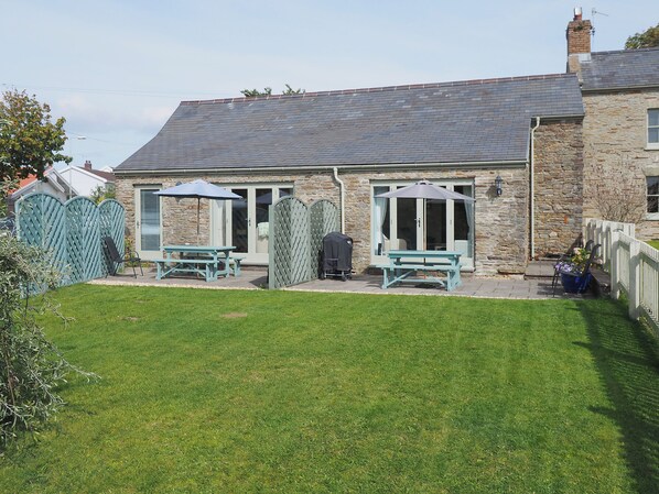 A stone house with a tiled roof, a fenced backyard, and two picnic tables with umbrellas.