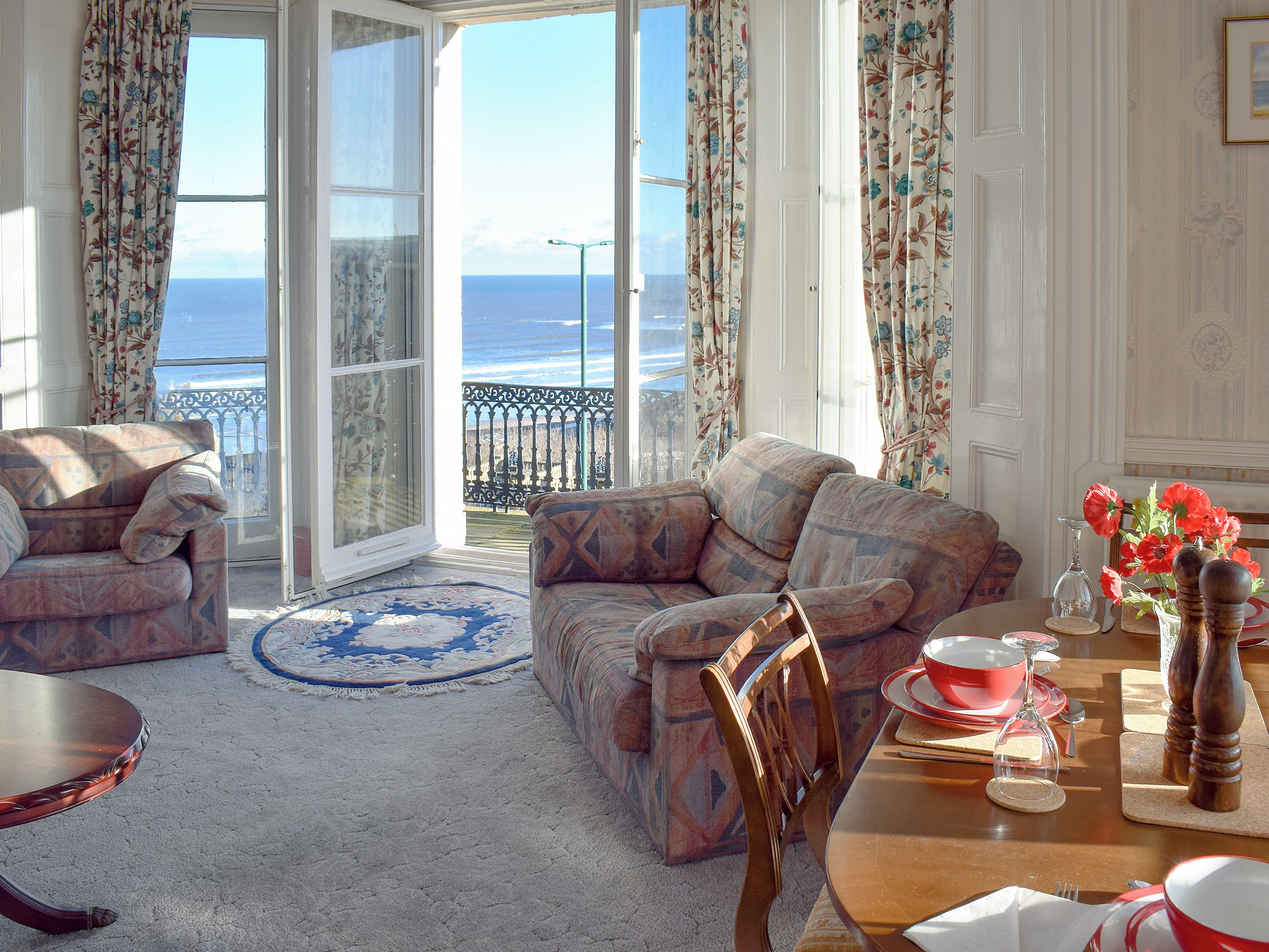 A room with a view of the sea, furnished with a sofa, armchair, and a dining table set for two.