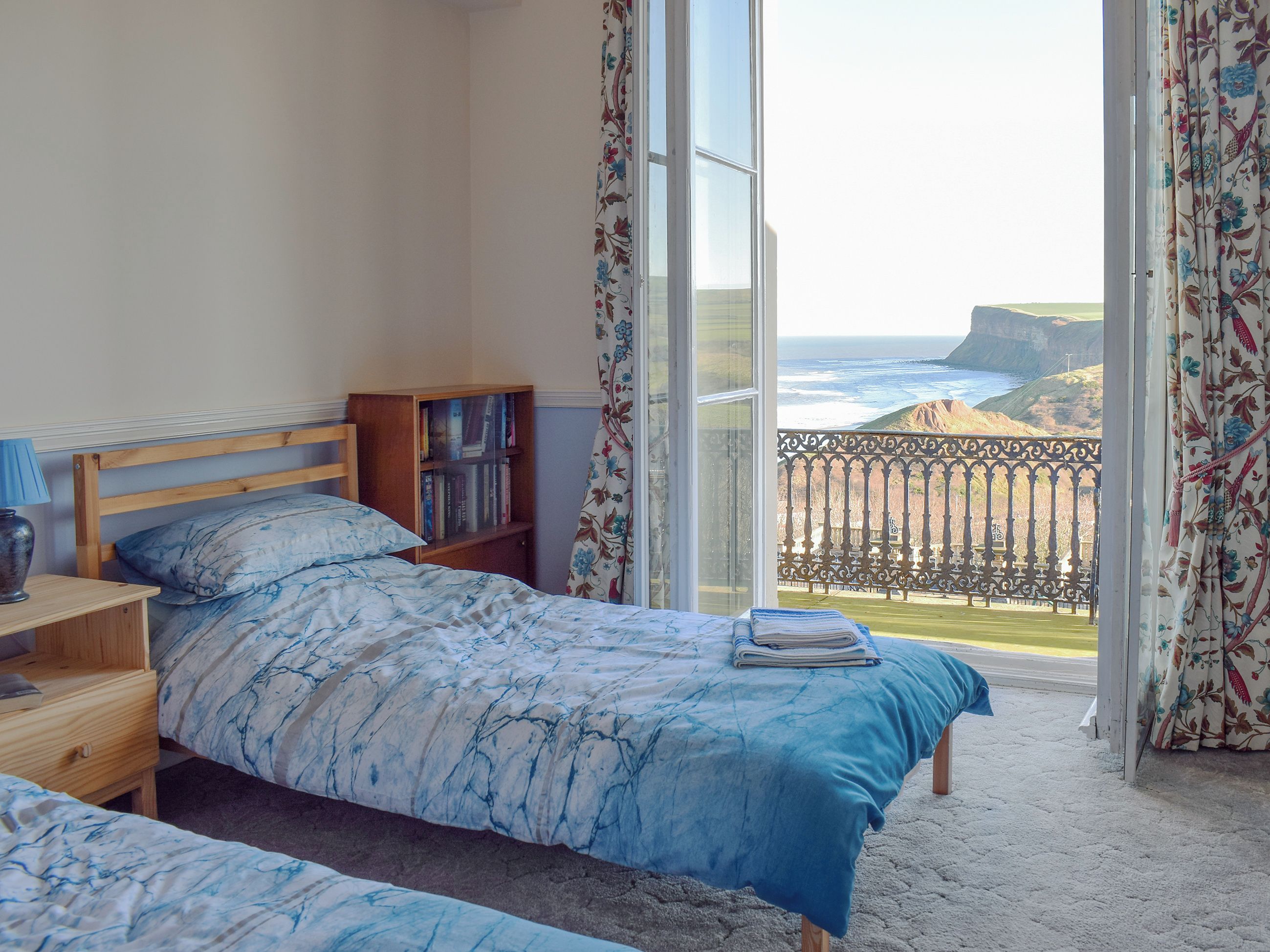 A bedroom with a bed, a bedside table, a bookshelf, and a view of the ocean through a balcony.