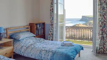 A bedroom with a bed, a bedside table, a bookshelf, and a view of the ocean through a balcony.