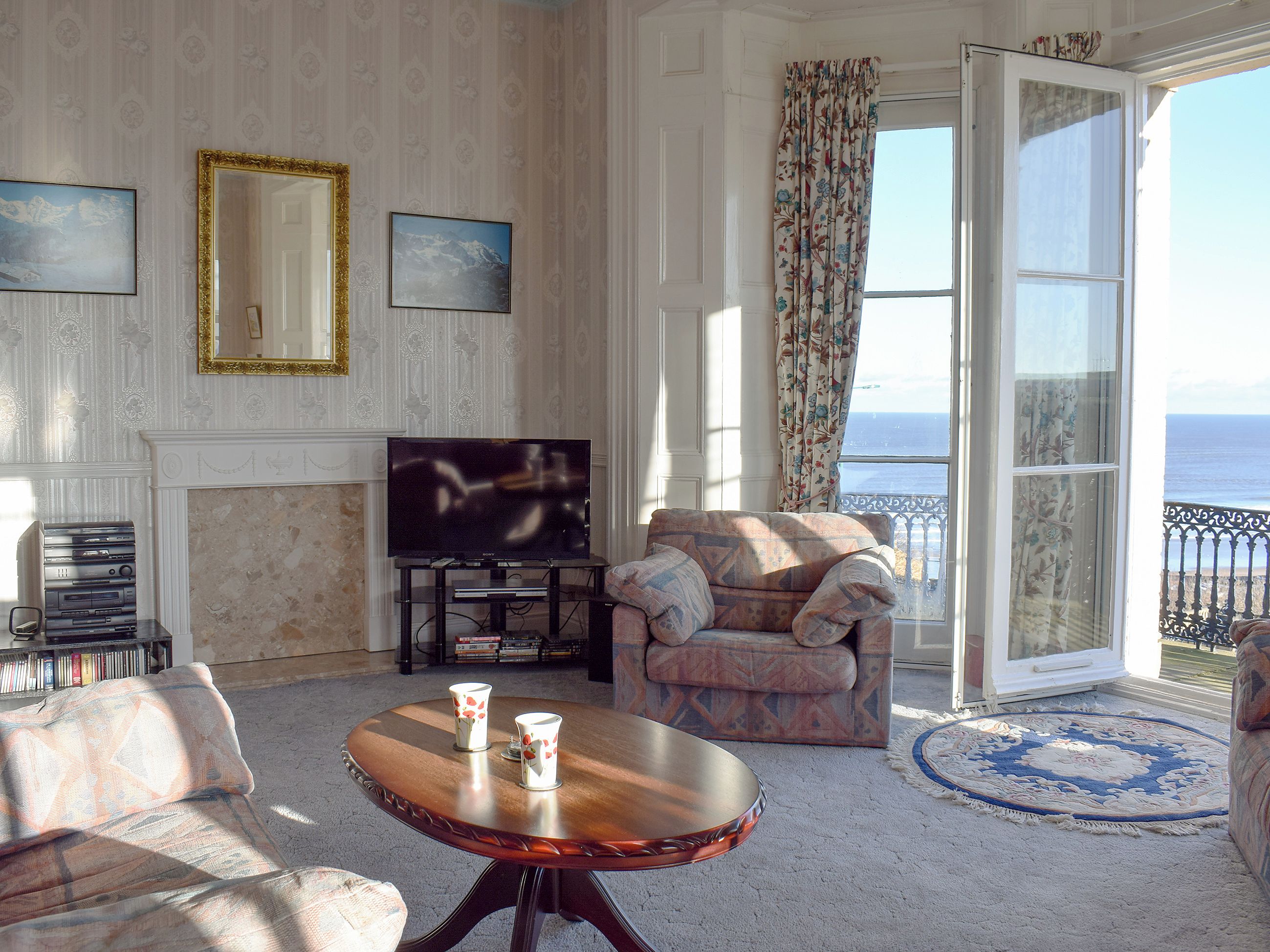 A living room with a television, a sofa, a coffee table, and a view of the sea through a large window.