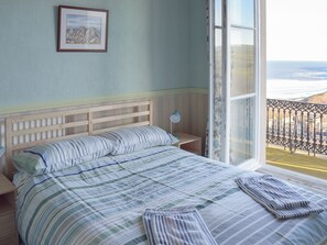 A bedroom with a bed, a nightstand, a lamp, and a window with a view of the beach.