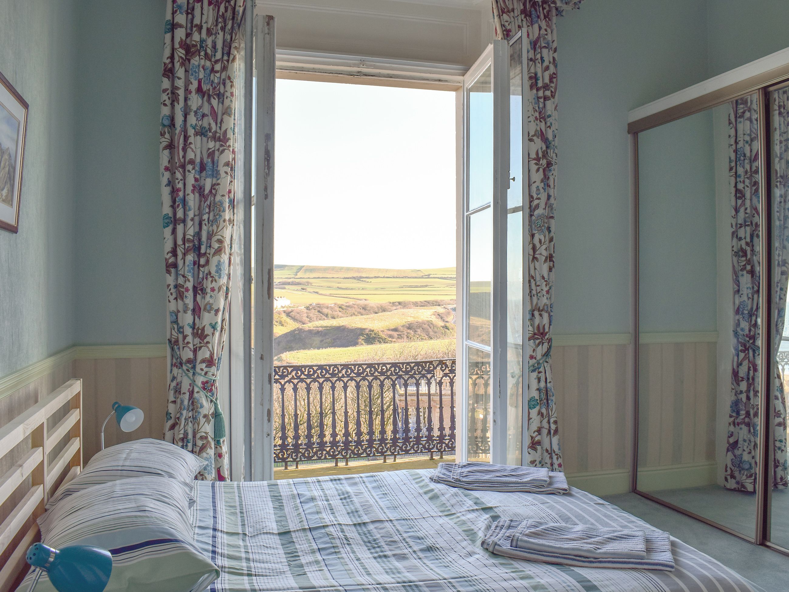 A bedroom with a bed, a bedside lamp, a mirror, and a view of the countryside through an open door.