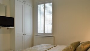 Un dormitorio con cama, televisor en un estante, ventana con persianas y radiador.