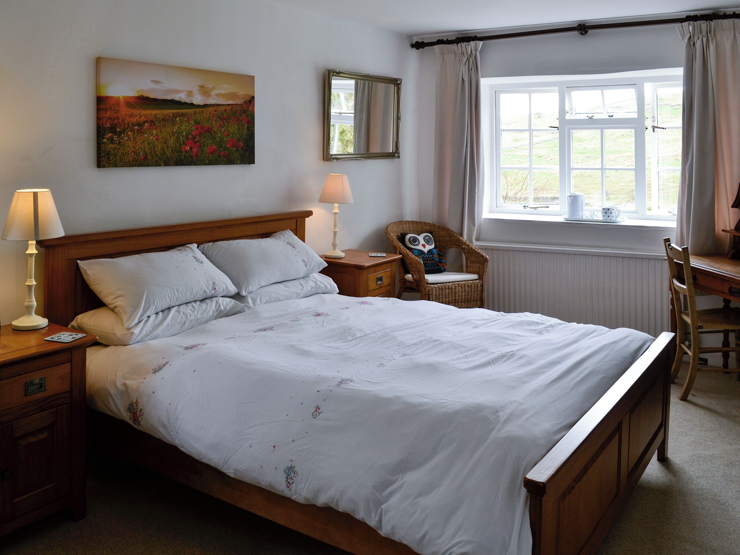 A bedroom with a wooden bed, two bedside tables with lamps, a wicker chair, a window with curtains, and a painting on the wall.
