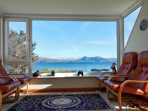 A room with a large window overlooking a body of water and mountains, two leather chairs, a patterned rug, and a small table with binoculars.