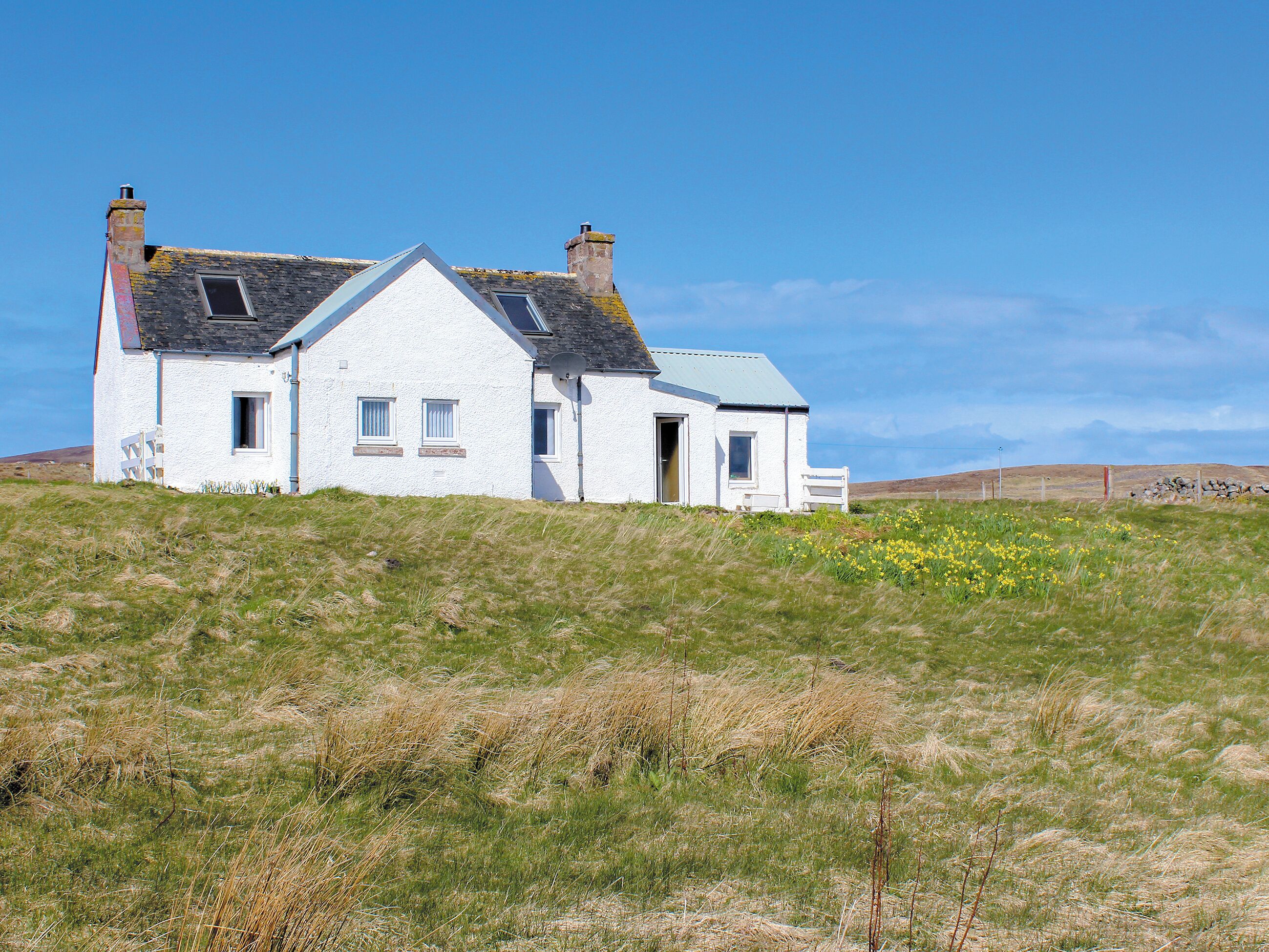 A white house with a gabled roof, situated on a grassy hill with a clear sky.
