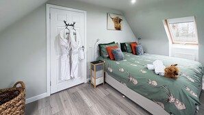 Un dormitorio con una cama, una mesita de noche de madera, una cesta de mimbre y una puerta con batas blancas colgadas en un perchero.