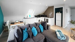 Una sala moderna con un sofá seccional gris, una pequeña mesa de comedor y una cocina con alacenas blancas y un backsplash de ladrillo.
