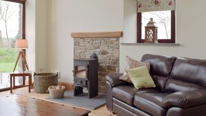 A living room with a stone fireplace, a leather sofa, a lamp, and a window with a view of trees.