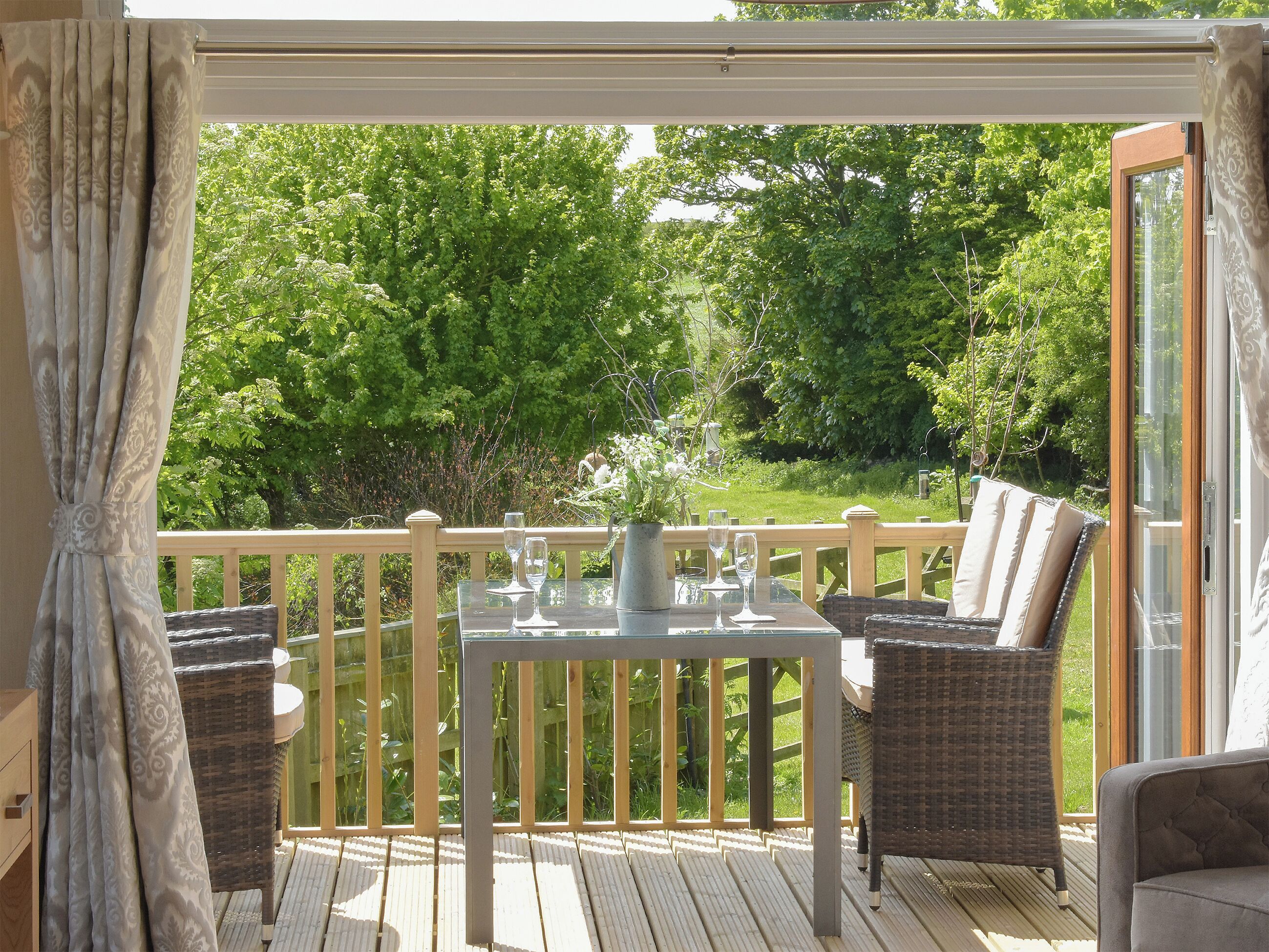 A covered outdoor seating area with a table and chairs, a view of a lush garden, and a wooden deck.