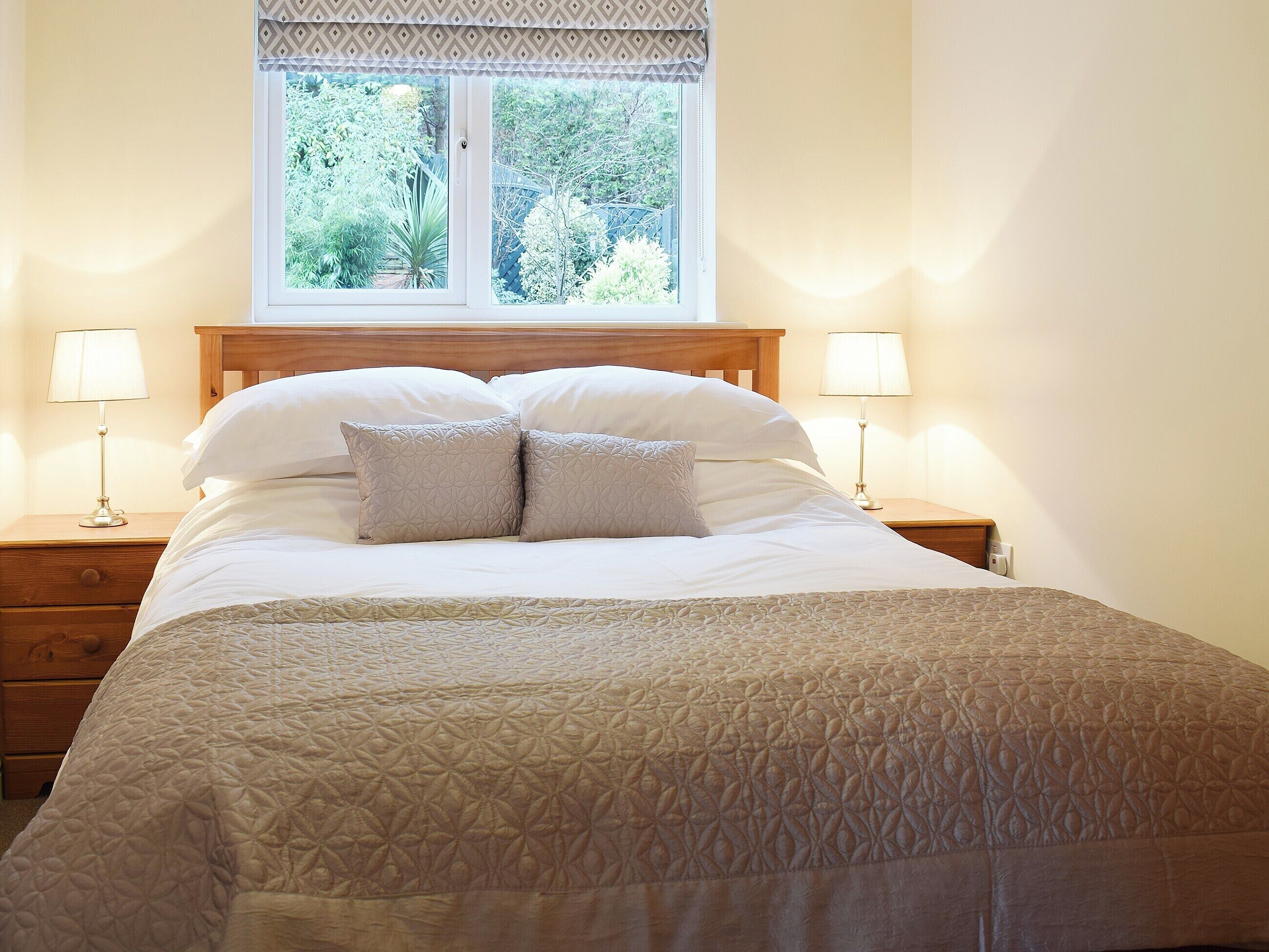 A neatly made bed with a patterned bedspread, two bedside lamps, and a window with a view of greenery.